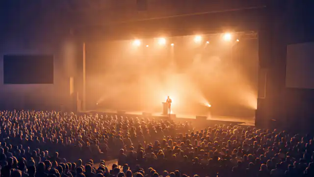 A packed auditorium from the back, stage lights raking through haze onto a lone speaker at a lectern.