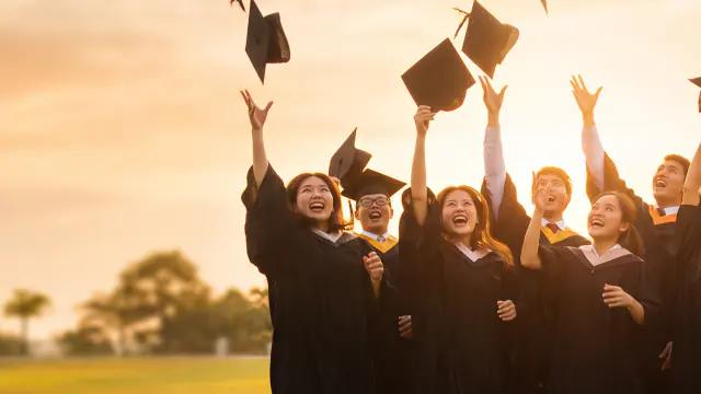 Graduates laughing as they throw their mortarboards into a gold evening sky.