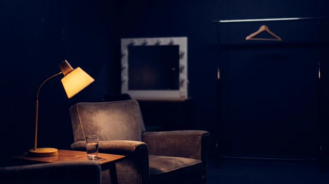 A backstage green room: one armchair, a lit lamp, a glass of water and a mirror with the bulbs off.