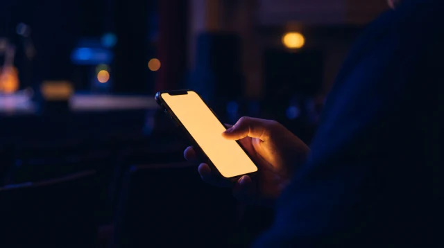 A hand holding a phone in a dark auditorium, its screen the brightest thing in the frame.